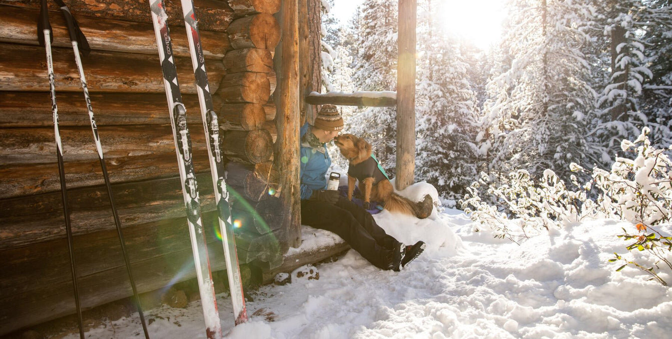 Dog licks humans face while sitting on step of nordic warming hut in the snow.
