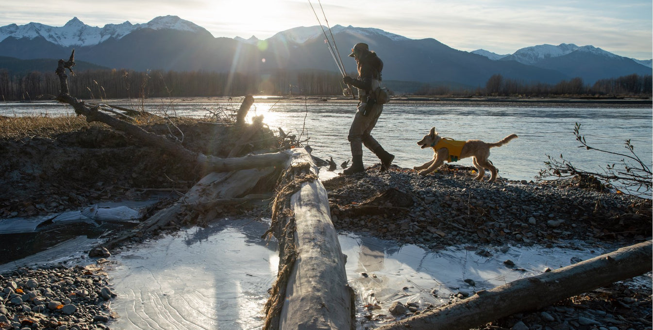 Chris carries fishing pole walking along river while dog runs behind in Float Coat.