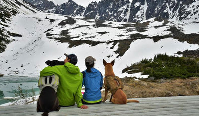 Laura, her partner and dogs sit on a dock looking out a snowy lake and mountains.