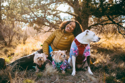 Alexis smiling laying on the ground in a meadow with her three dogs in matching blossom pattern dog sweaters.
