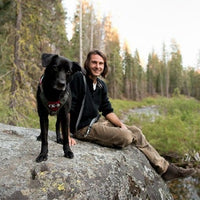 Black lab and man sit on rock in the woods by a river.