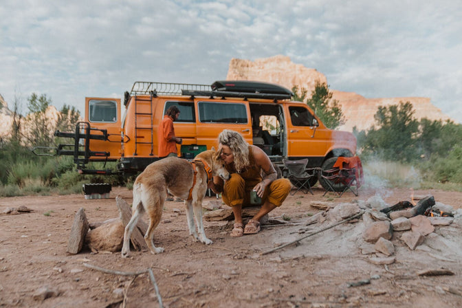 Woman in orange overalls kisses dog in orange front range harness while they sit in front of orange van in desert!